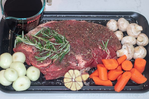 uncooked chuck roast with carrots, onions, mushrooms, garlic, herbs and wine on a platter