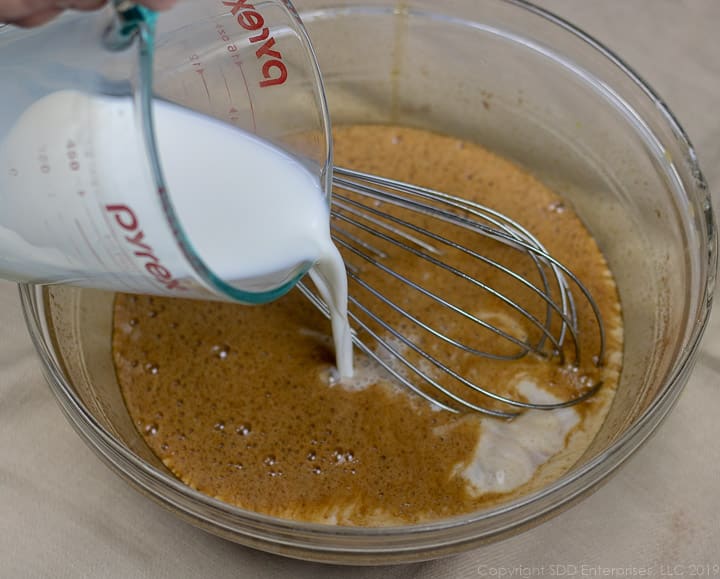 pouring whole milk into a clear glass bowl full of egg and spice mixture for bread pudding