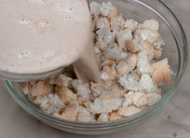 pouring custard mix over french bread in a clear glass bowl for bread pudding