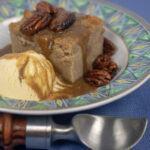 bread pudding with cane syrup rum sauce and glazed pecans with a scoop of vanilla ice cream in a bowl with green trim and an ice cream scooper