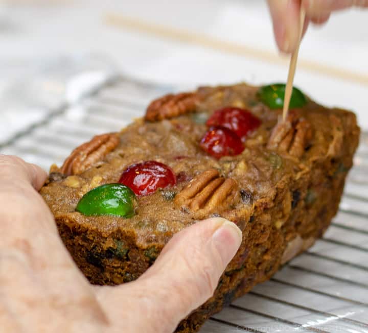 A hand poking the top of the fruitcake on a cooling rack with a toothpick.