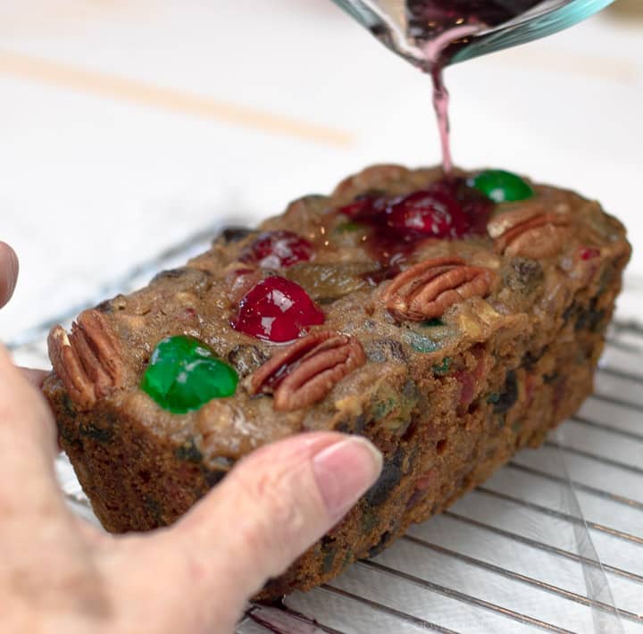 Pouring brandy from a measuring cup on the top of a fruitcake on a cooling rack>