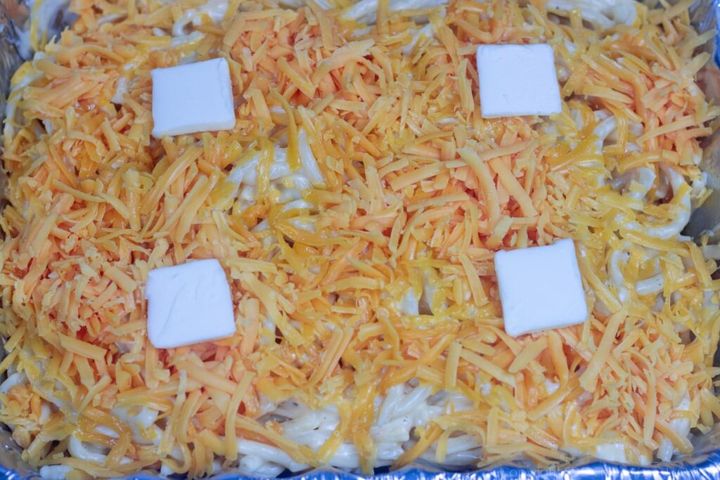 noodles in cheese sauce covered with grated cheese and four tabs of butter for macaroni and cheese ready for the oven