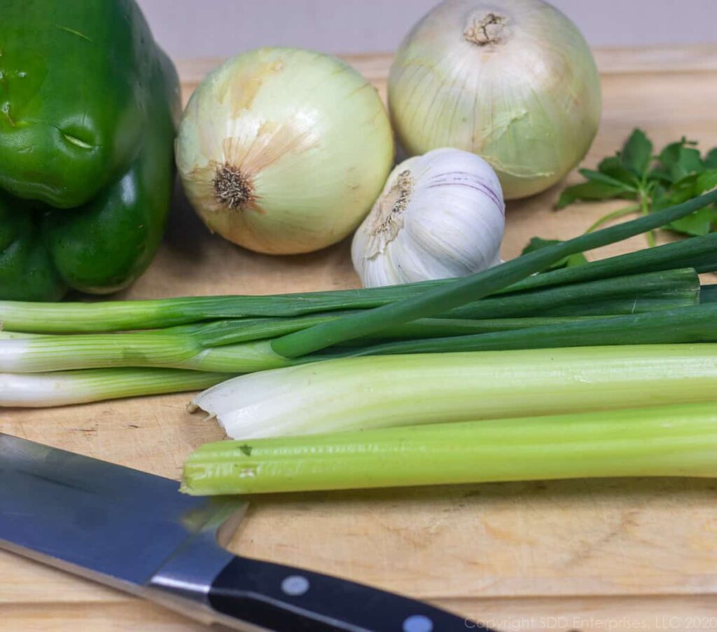 yellow onions, green onions, bell peppers, celery and garlic on a cutting board with a knife