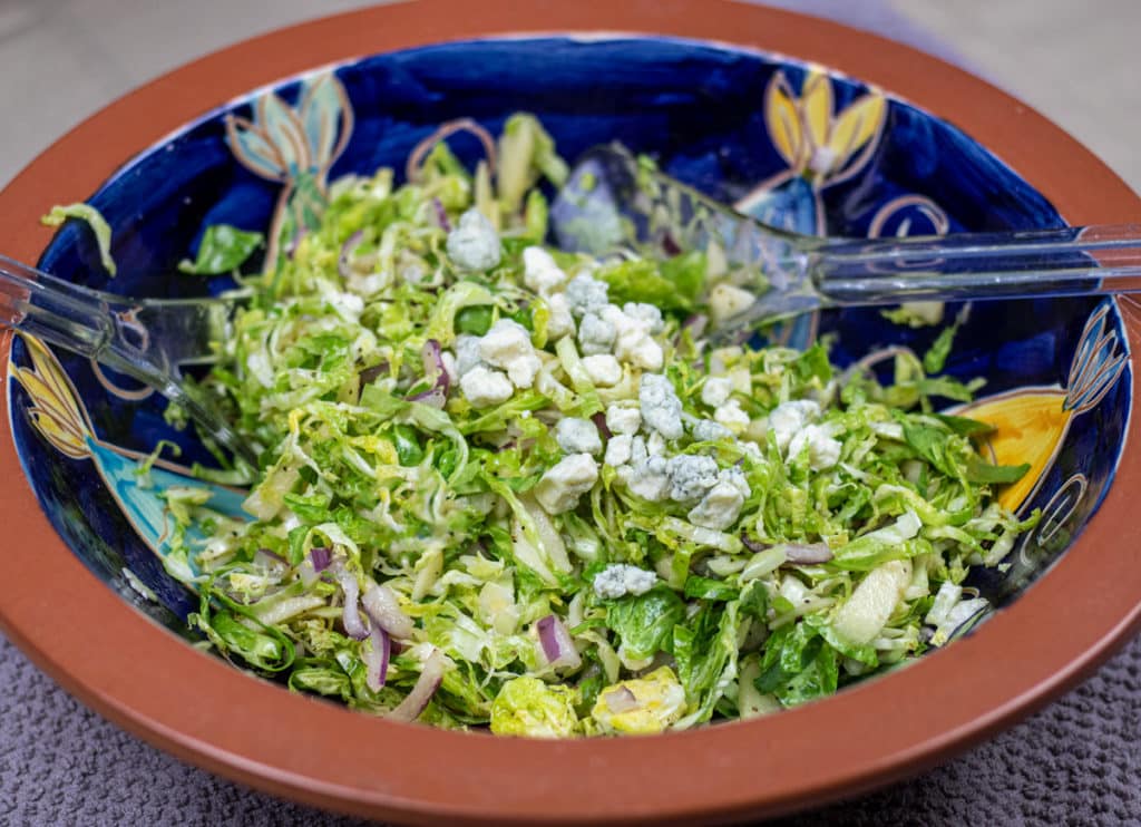 brussel sprout slaw with blue cheese garnish in a brown and blue bowl with salad tongs