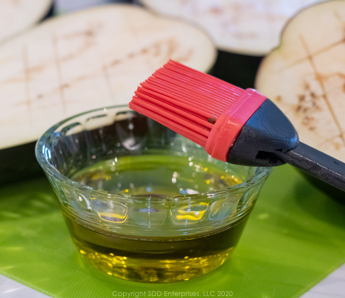 eggplant sliced in half and cross-hatched with a paring knife and a small bowl of olive oil with a silicon brush