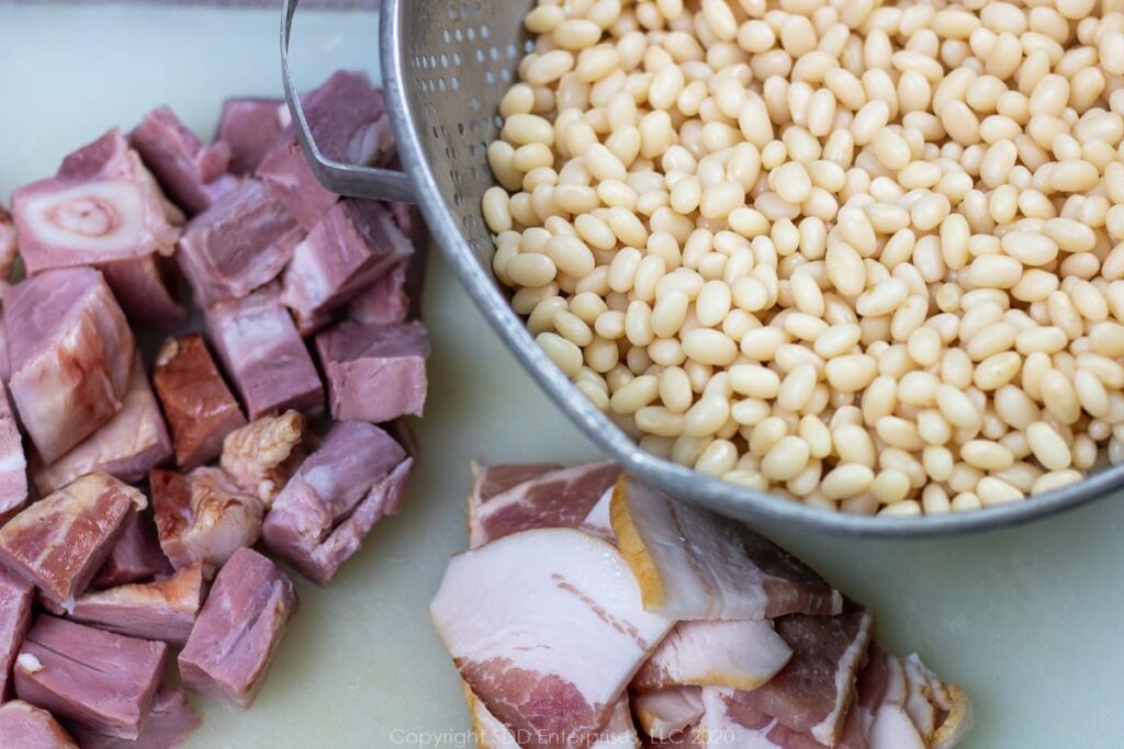 dry white beans in a colander, cubed ham and sliced bacon on a cutting board