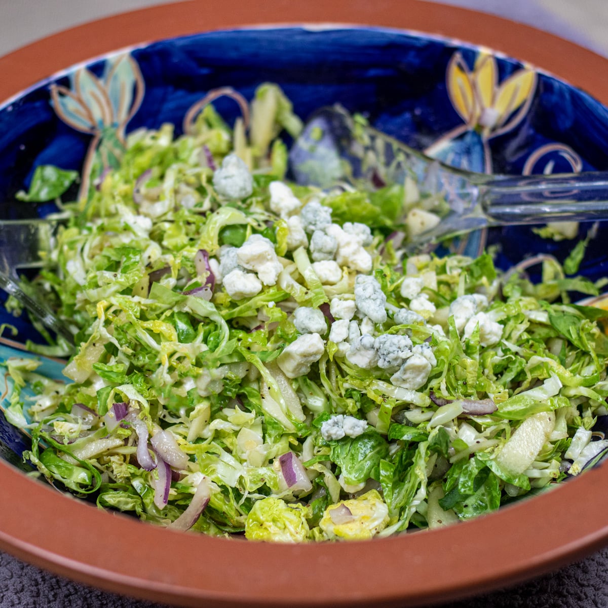 brussels sprouts coleslaw with blue cheese in a blue and brown bowl with salad tings