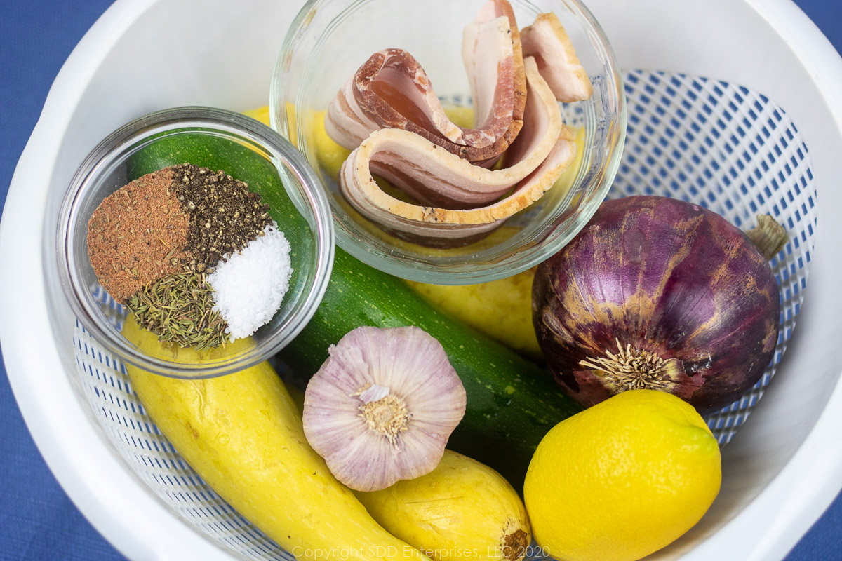 fresh yellow squash, zucchini, garlic, red onion, bacon and herbs and spices in a white colander. 