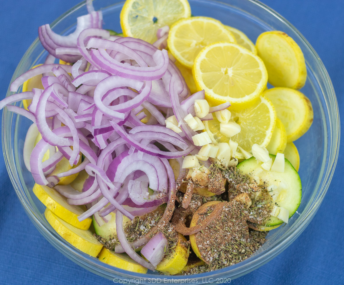 sliced red onion, lemons, chopped garlic and spices added to slice squash and zucchini in a glass bowl