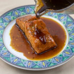 roasted salmon filet on a white plate with blue trim with cane syrup sauce being poured from a sauce pan