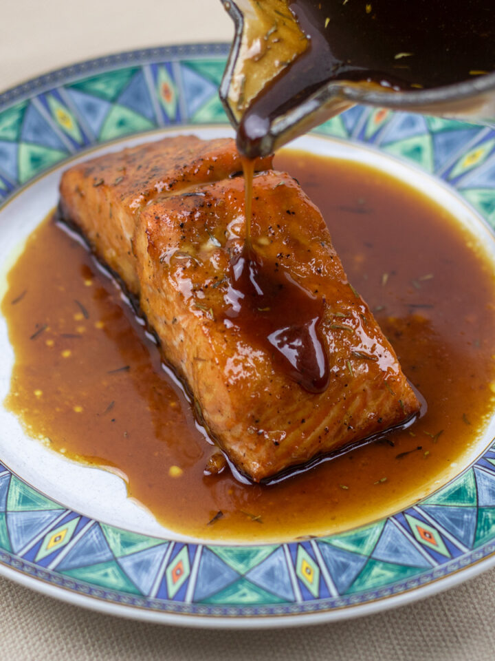 roasted salmon filet on a white plate with blue trim with cane syrup sauce being poured from a sauce pan