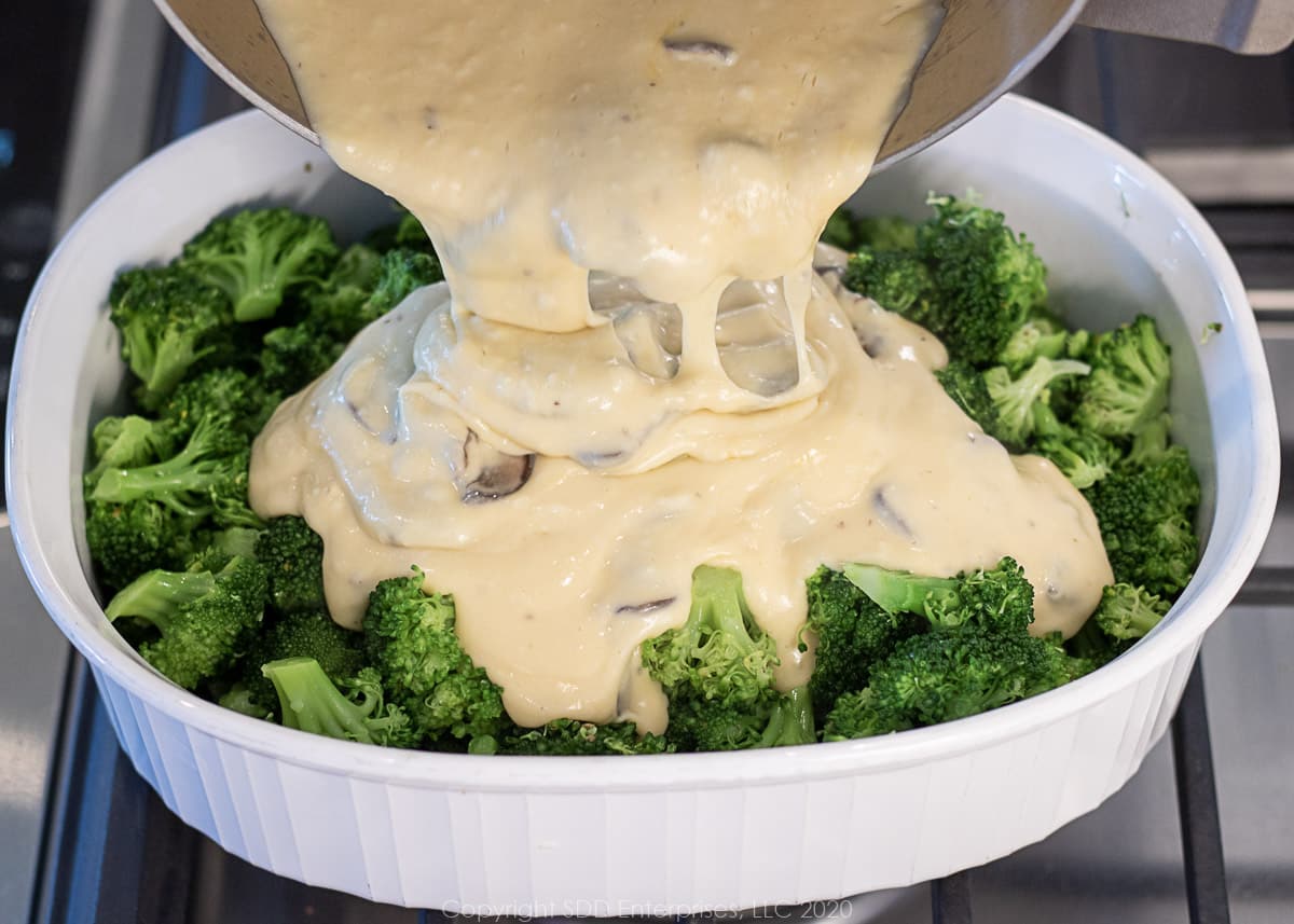 pouring cheese sauce over broccoli florets in a baking dish