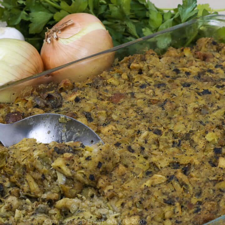 oyster artichoke dressing in a baking dish with a serving spoon and onions, garlic and parsley garnish