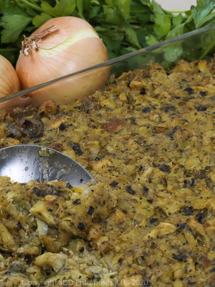 oyster artichoke dressing in a baking dish with a serving spoon and onions, garlic and parsley garnish