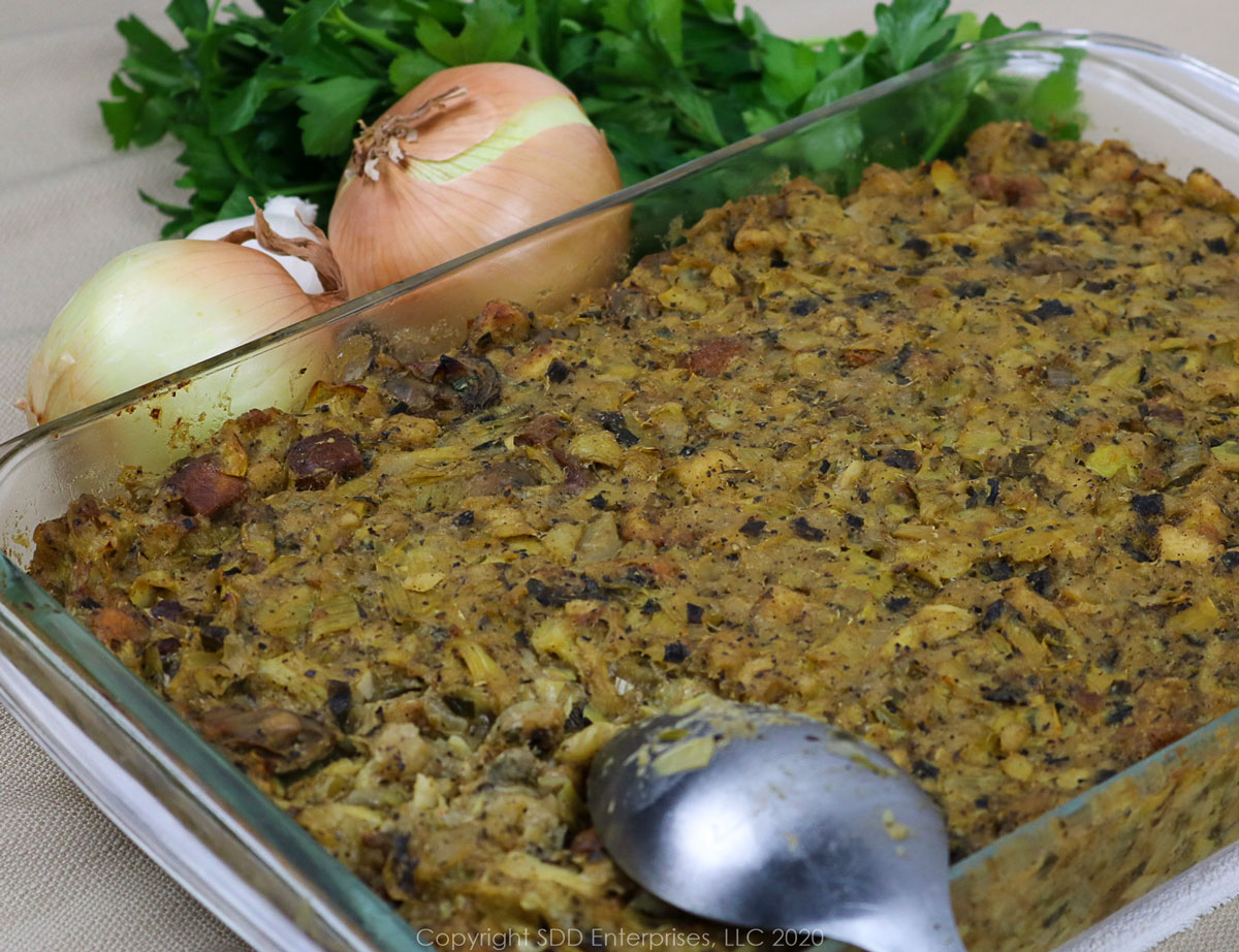 oyster artichoke dressing in a baking dish with a serving spoon and onions, garlic and parsley garnish