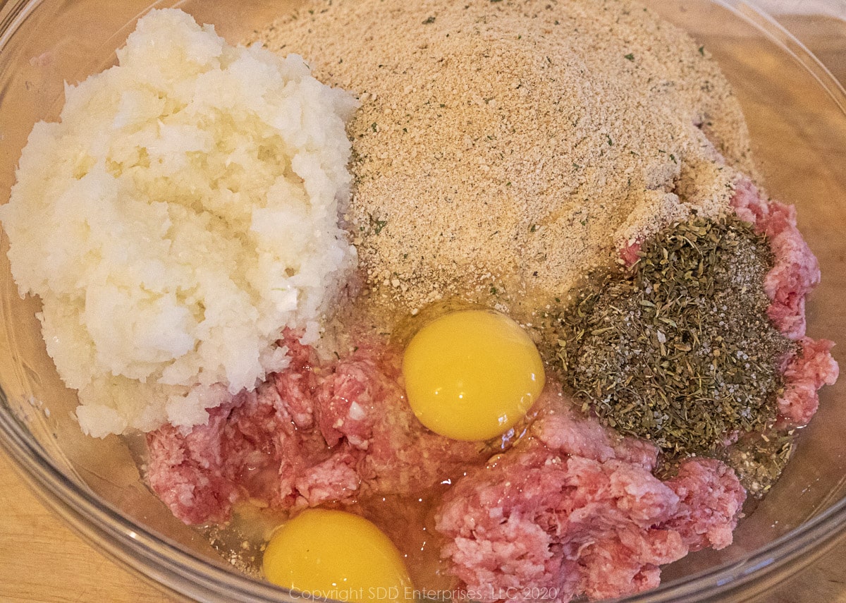 onions, garlic, eggs, breadcrumbs and spices added to ground meats in a glass bowl