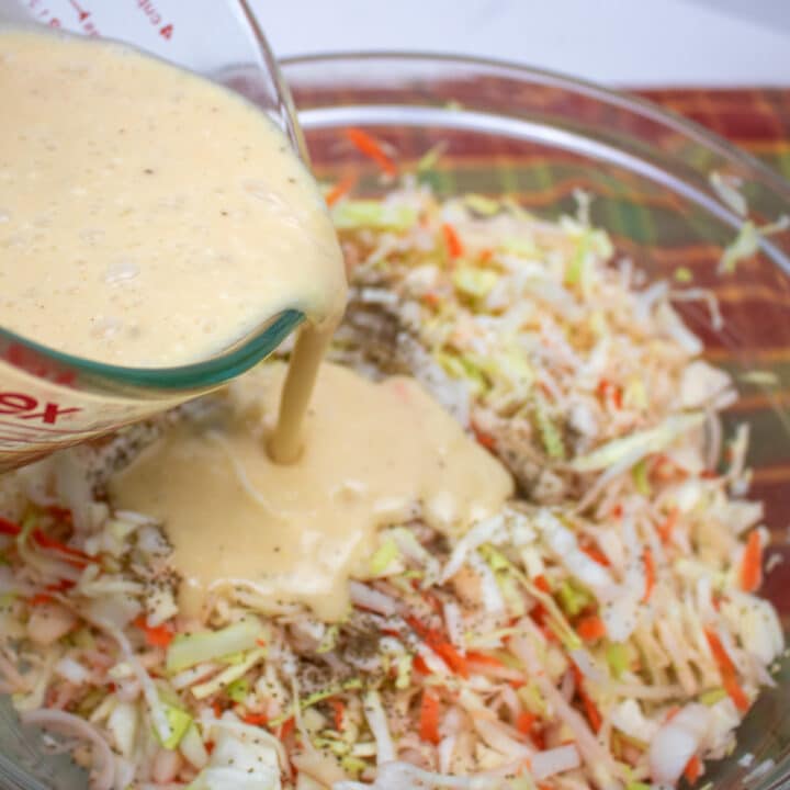 creamy dressing being poured on shredded cabbage, apples, carrots and onions in a glass bowl