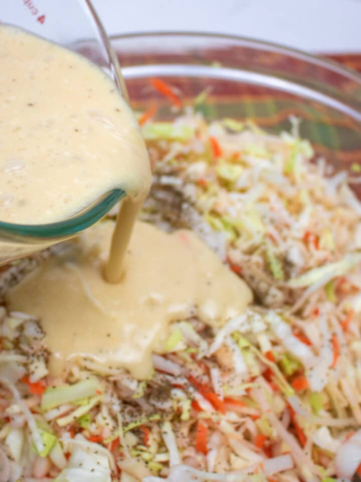 creamy dressing being poured on shredded cabbage, apples, carrots and onions in a glass bowl