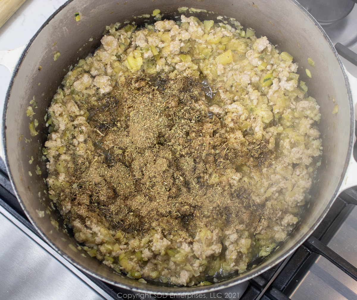 herbs and spices being added to simmering vegetables and pork in a Dutch oven