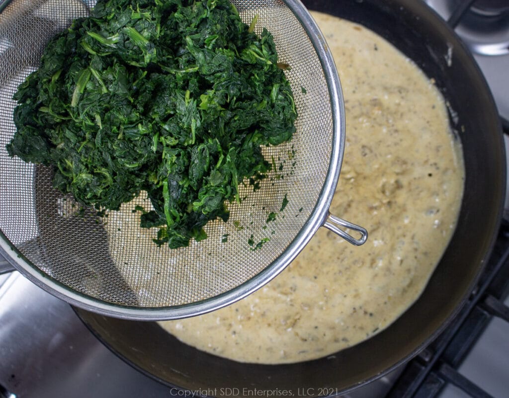Adding drained spinach to cream sauce in a sauce pan.