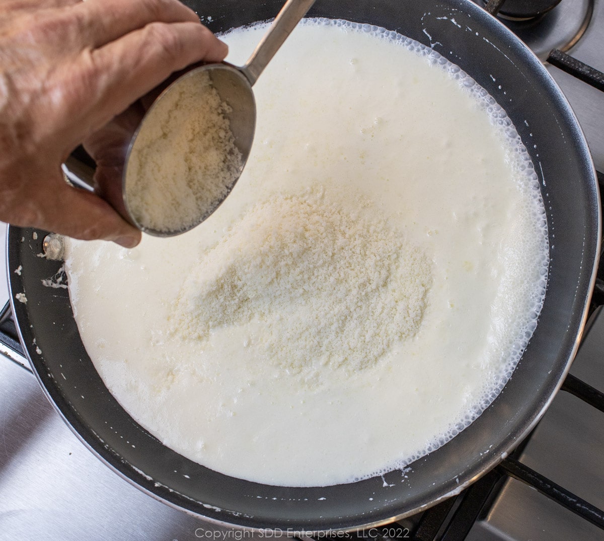 grated cheese being added to heavy cream in a sauté pan