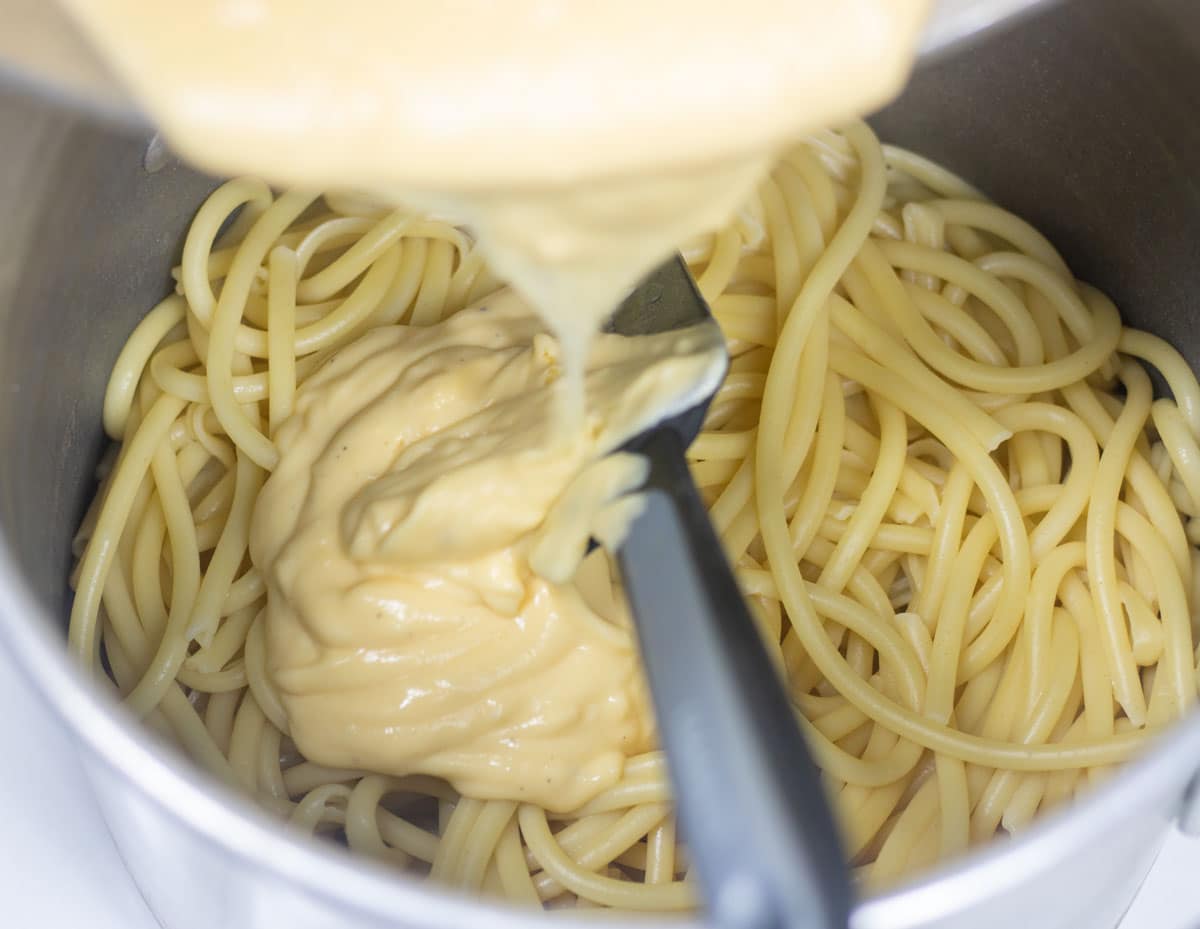 cheese sauce being poured onto cooked pasta in a stock pot