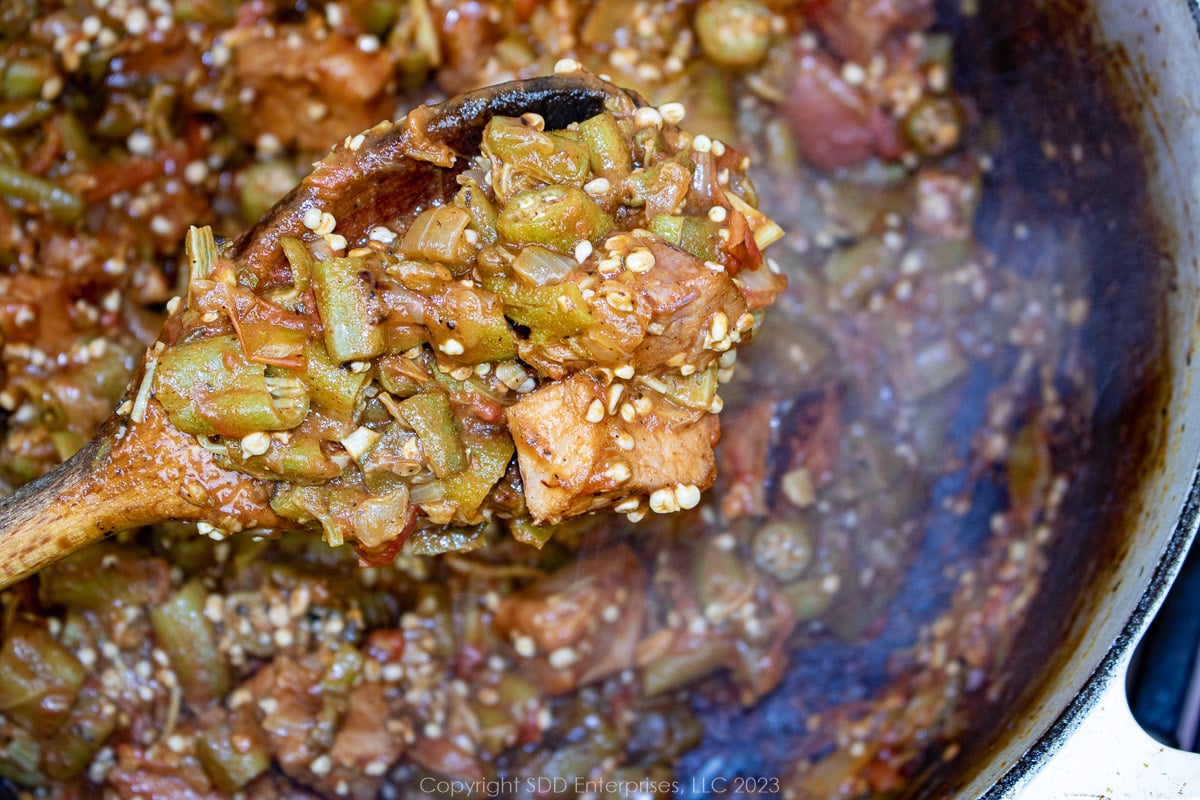 smothered okra and tomatoes with ham on a wooden spoon over a Dutch oven