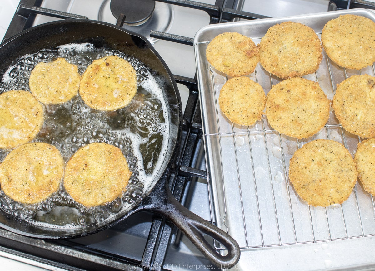 green tomatoes frying in a cast iron skillet and draining on a cooling rack