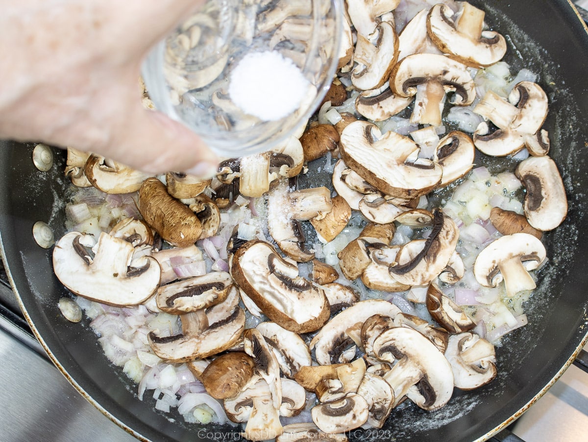 salt being added to mushrooms and shallots in a skillet