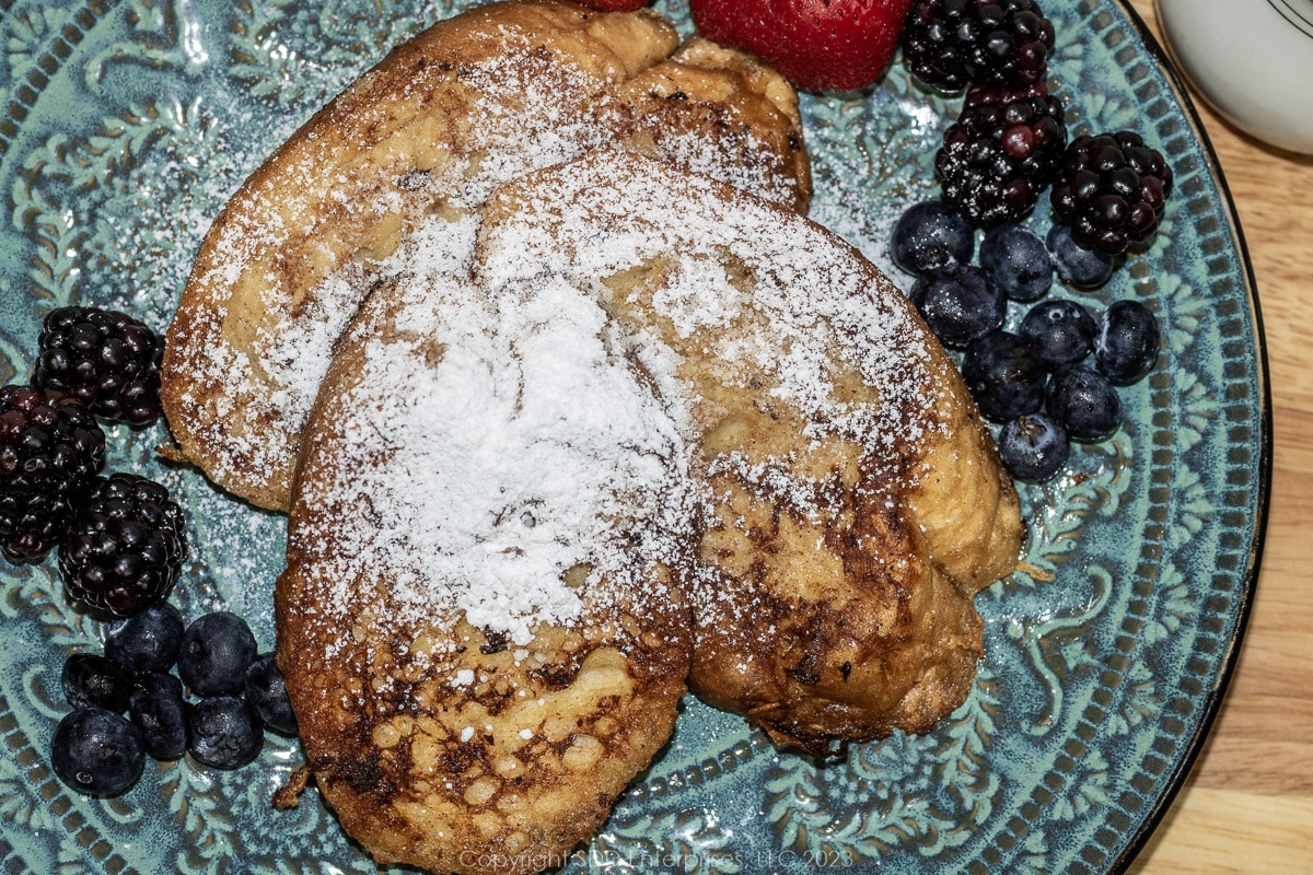 Pain Perdu or French Toast on a blue green plate with powdered sugar and fruit garnish.