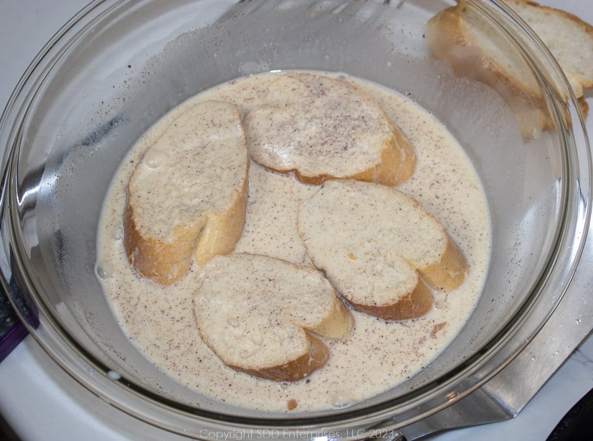 Slices of French bread soaking in custard in a glass bowl.