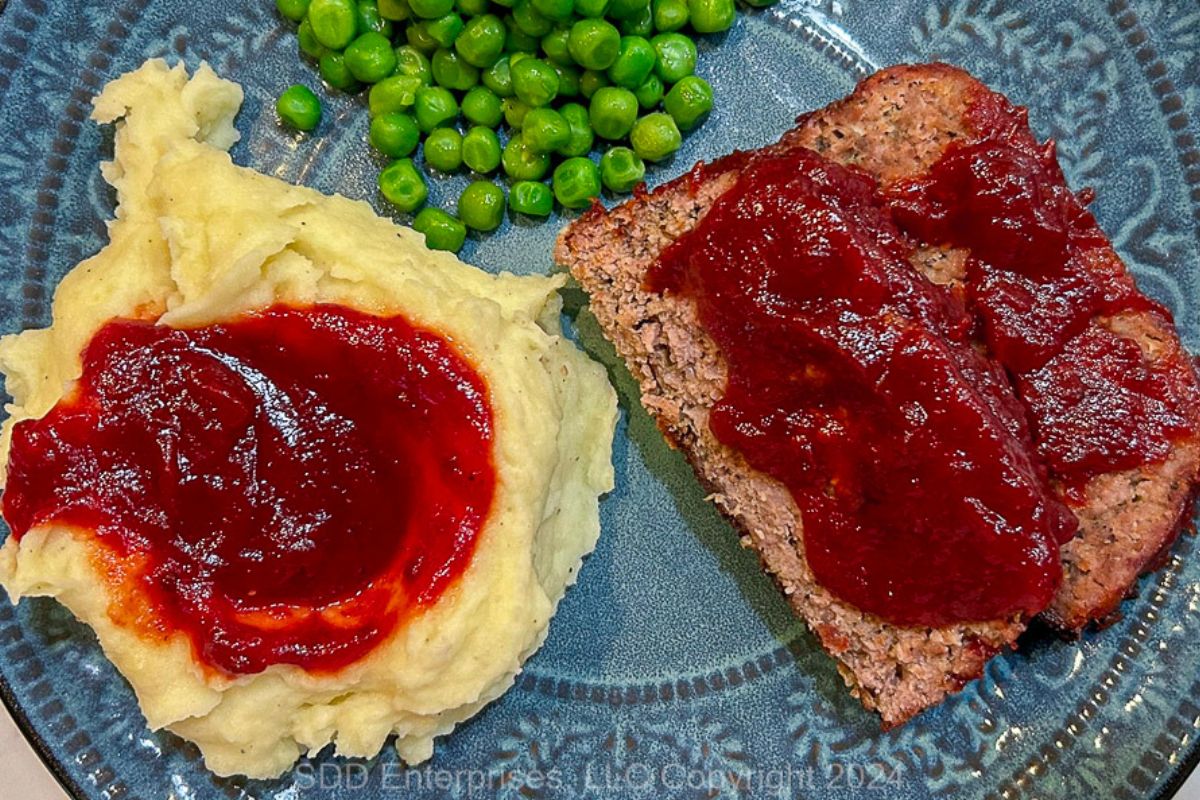 Sliced meatloaf with sauce with mashed potatoes and peas on a blue-green plate.