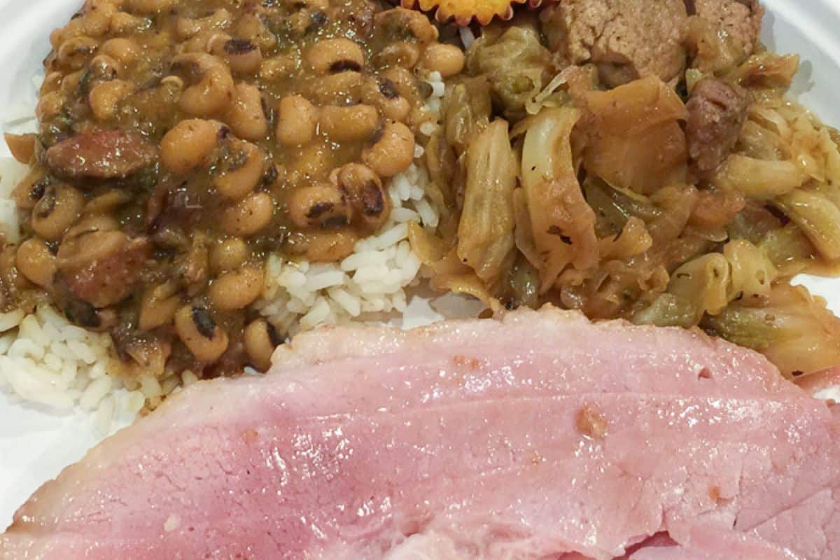 Black-eyed Peas over rice served with smothered cabbage and baked ham on a white plate.