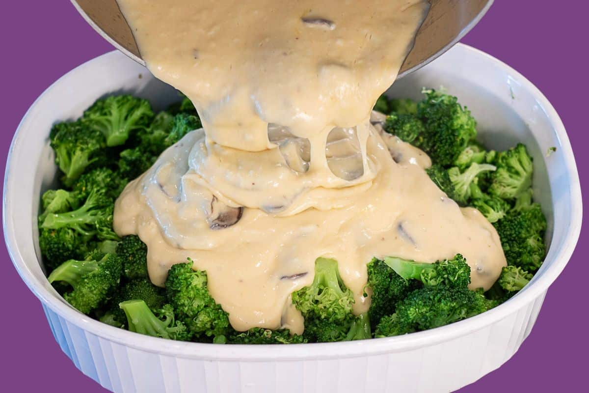 Rich béchamel sauce being poured from a saucepan over fresh broccoli florets in a casserole dish.