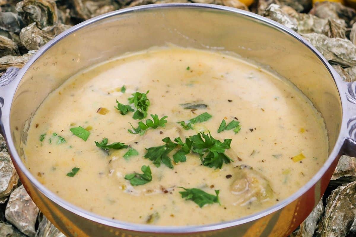 Oyster Soup with garnish in a silver serving bowl overlaid on oyster shells.